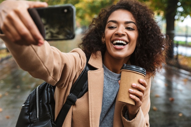 아름 다운 젊은 아프리카 여자는 selfie를 복용, 배낭을 들고 야외 공원에서 산책하는 코트를 입고