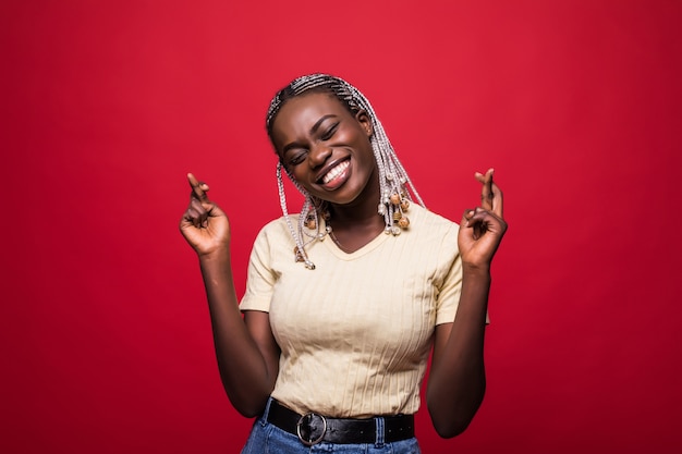 Beautiful young Afro American woman is holding fingers crossed and smiling, isolated on red background
