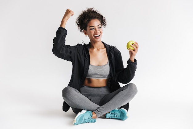 Bella giovane incredibile fitness sportivo donna africana mangia mela isolato sul muro bianco.