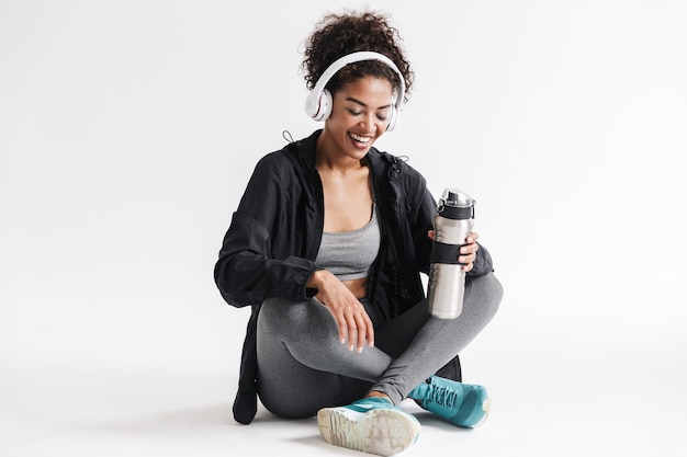 beautiful young amazing sports fitness african woman listening music in headphones isolated over white wall drinking water.