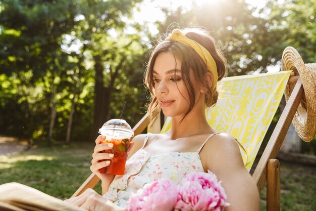 緑の公園でジュースを飲む美しい若い驚くべき女性は、本を読んでサンベッドに横たわっています。