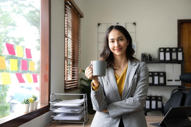 Bella giovane donna di affari asiatica che sorride tenendo una tazza di caffè e un computer portatile che lavorano all'ufficio