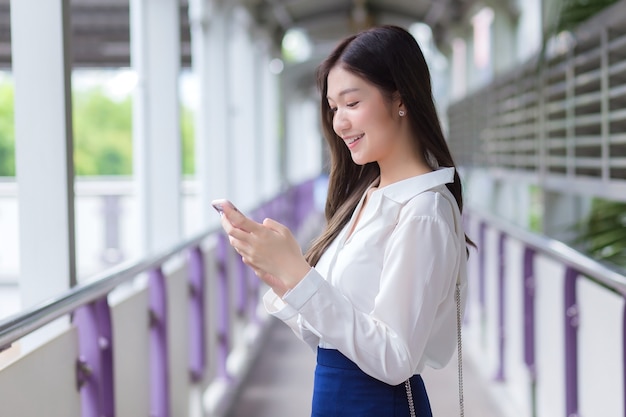 Beautiful young Asian businesswoman stands on flyover of skytrain outdoors in town while uses her smartphone to send messages to office.