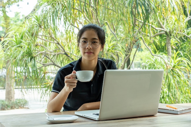 カフェでテーブルの上のノートパソコンとコーヒーを保持している美しい若いアジアの女の子。