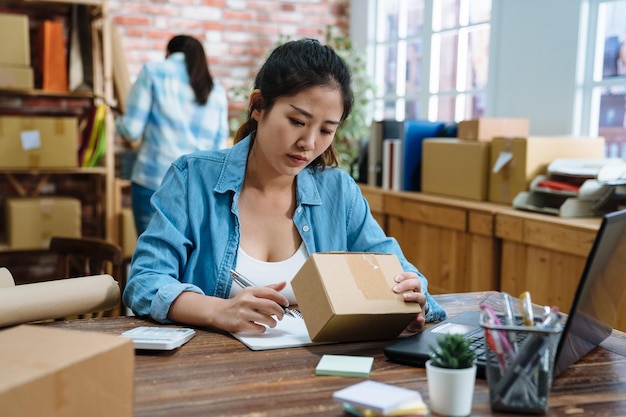 beautiful young asian girl works at home office. two women friends own online business small shop prepare goods to customers in studio. female freelancers in warehouse doing ecommerce sending parcel.