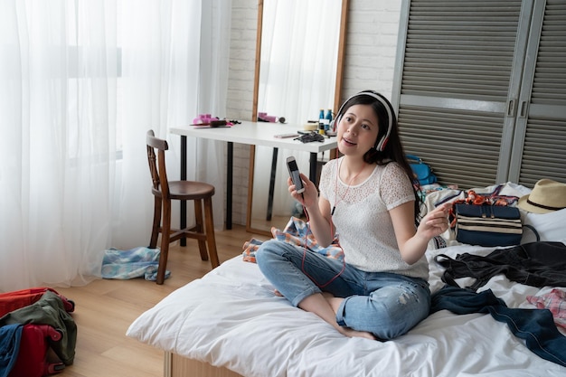 Bella giovane donna giapponese asiatica seduta sul letto nella camera da letto di casa utilizzando il cellulare ascolta musica scarica online con le cuffie. femmina con un sorriso perfetto e godendosi la melodia che canta felice