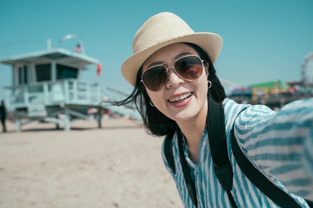 Beautiful young asian korean woman taking selfie on sandy beach by lifeguard tower. charming girl in sunglasses and straw hat face camera and smiling. female traveler enjoy sunshine under blue sky.