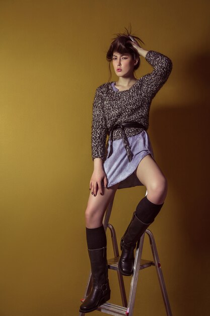 Beautiful young asian model in blue denim skirt and black blouse and fashion makeup posing sitting on stepladder and looking at camera. Studio shot. High-fashion photo.