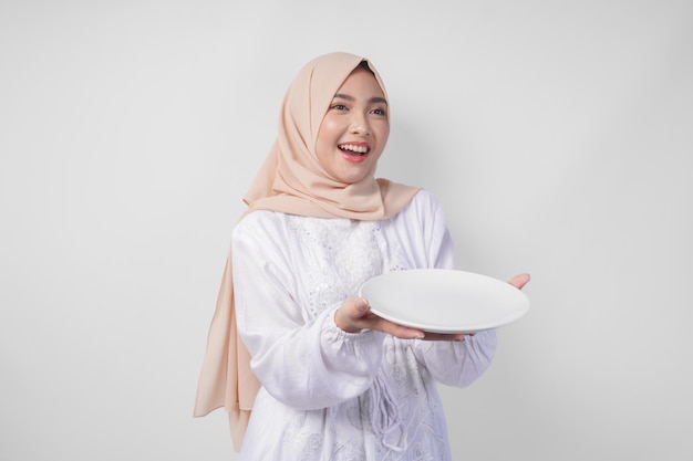 Foto bella giovane musulmana asiatica in hijab presenta un piatto vuoto con uno spazio di copia sopra che mostra il menu del cibo per l'iftar