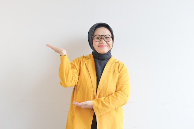 Beautiful young Asian Muslim woman wearing glasses and yellow blazer showing palms while smiling