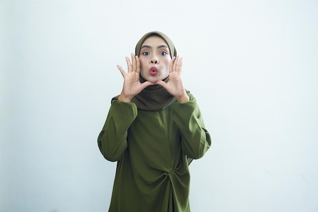 Beautiful young asian muslim woman wearing green dress and hijab screaming with hands beside mouth