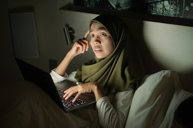 beautiful young Asian Muslim woman wearing headscarf while lying in bed and using laptop at night