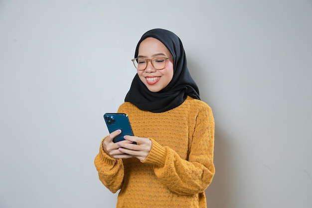 Красивая молодая азиатская мусульманка в оранжевом свитере и очках при использовании мобильного телефона
