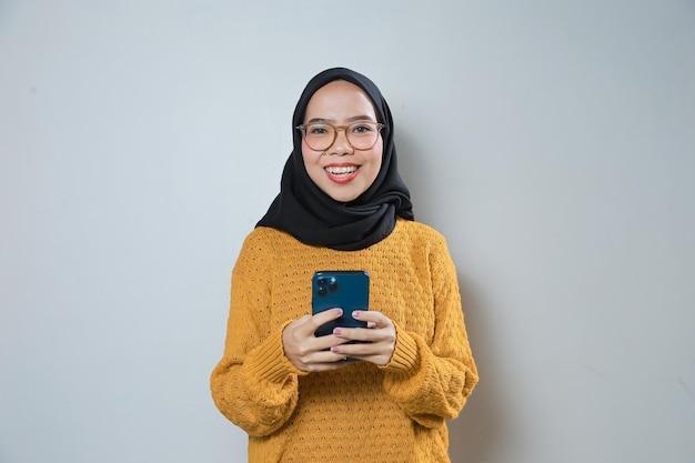 Beautiful young asian muslim woman wearing orange sweater and glasses while using mobile phone