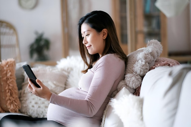 Beautiful young Asian pregnant woman using smartphone while relaxing on sofa in the living room enjoying at cozy home Wellbeing Pregnancy lifestyle