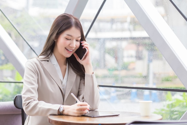 Beautiful young Asian professional business woman in a cream-colored suit holds a smartphone to talk, chat and record information happily on a tablet while in a coffee shop.