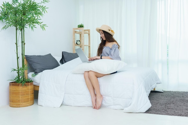 Beautiful young Asian woman in blue and white striped casual dress smiling and using laptop in the bedroom