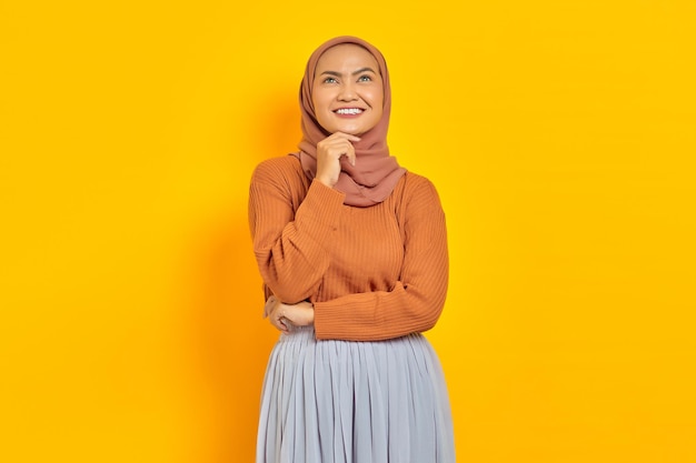 Beautiful young asian woman in brown sweater and hijab smiling
looking up with hand on chin, having good idea isolated over yellow
background. people islam religious concept