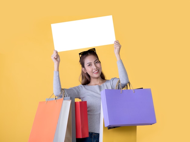 Beautiful young Asian woman carrying shopping bags and holging white banner with copy space isolated on yellow background