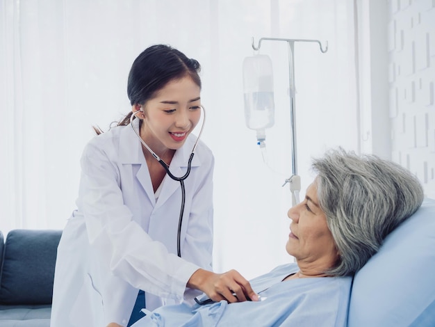 A beautiful young Asian woman doctor in white suit smiled while using stethoscope to examine listen to heartbeat of elderly old female patient in blue dress who was lying on bed in hospital room