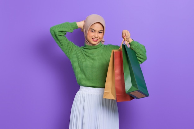 Beautiful young asian woman in green sweater holding shopping\
bags and scratching head with hand isolated over purple\
background