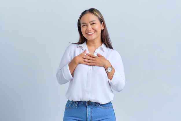 Beautiful young Asian woman holding hands on heart looking pleased and happy isolated over white background