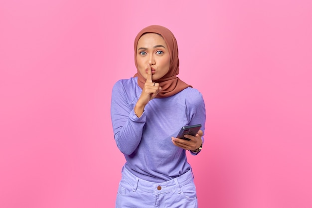 Beautiful young Asian woman holding mobile phone and makes silence gesture on pink background