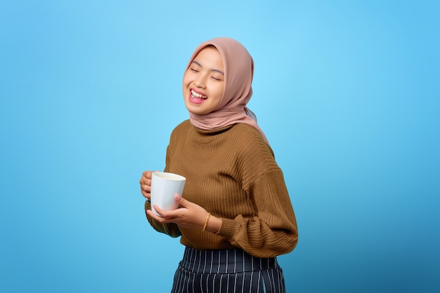 Beautiful young Asian woman holding mug and drinking tea on blue background