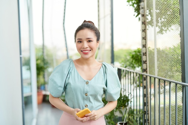 Beautiful young Asian woman holding phone and standing on the balcony