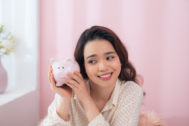 Beautiful young Asian woman holding piggy bank over pink wall.