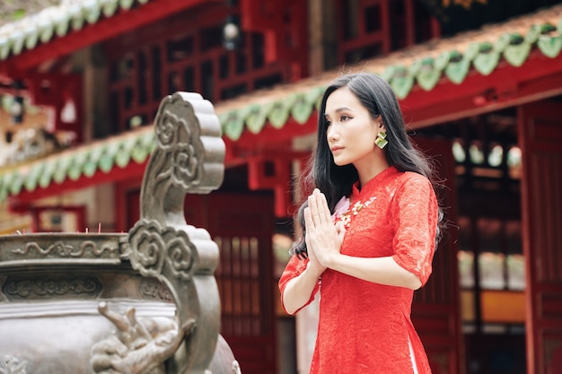 Beautiful young Asian woman in lace red dress making pray gesture when standing at ancient bronze urn with incense sticks at Buddhist temple