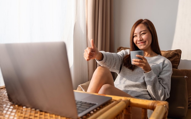 A beautiful young asian woman making thumb up hands sign while using laptop computer for video call at home