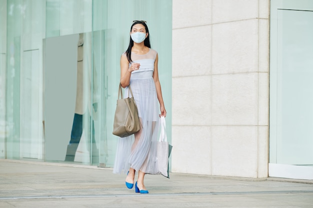 Beautiful young Asian woman in medical mask walking down empty city street