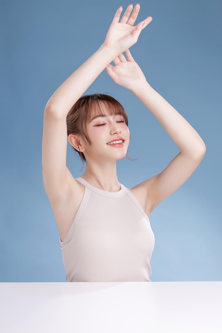 美麗年輕的亞洲女性模特兒，有瀏海和自然的臉部妝容，乾淨清新的皮膚，在孤立的藍色背景上展示腋下。可愛的女孩肖像，面部護理，身體護理，美容和水療