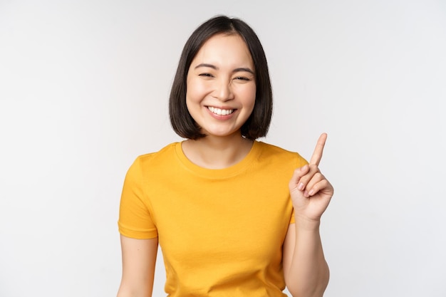 Beautiful young asian woman pointing finger up smiling and looking amused at camera showing advertisement announcement on top white background