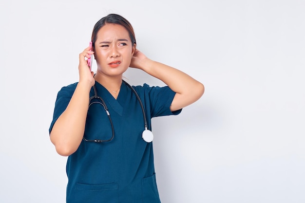 白い背景に分離された混乱した顔で脇を見て携帯電話で話している青い制服を着て働く美しい若いアジア女性専門の看護師医療医学の概念