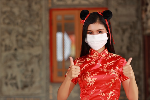 赤いチャイナドレスの美しい若いアジアの女性伝統的なチャイナドレスは、親指を上または二重のように示すジェスチャーでヘルスケア用の保護マスクを着用します（新しい通常および中国の旧正月の概念）