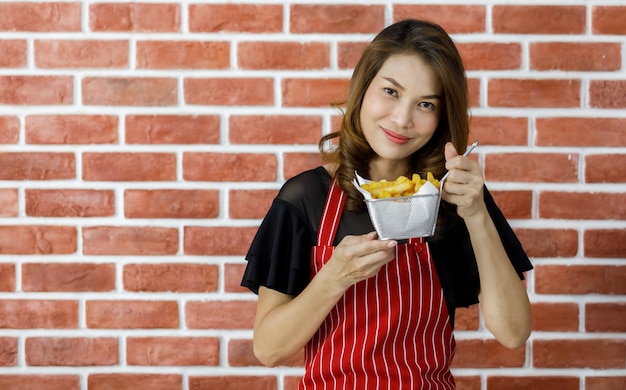 Beautiful young Asian woman in red striped apron holding sieve of tasty fried potatoes near brick wall and proudly show it with smile as satisfying her cooking skill and persuade eating delicious food