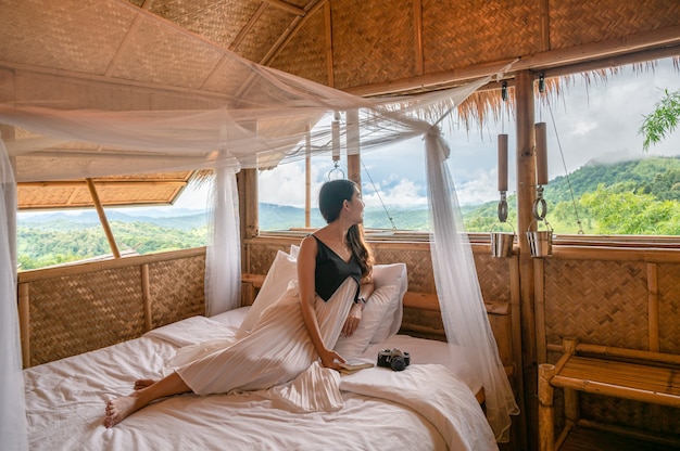 Beautiful young asian woman resting and looking at outside on the bed in wooden cottage among the mountain on vacation