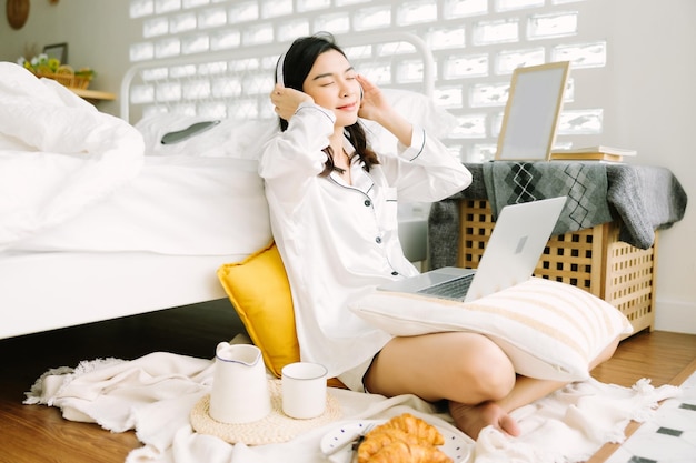 Beautiful young Asian woman sitting next to bed using laptop and breakfast in the morning at home