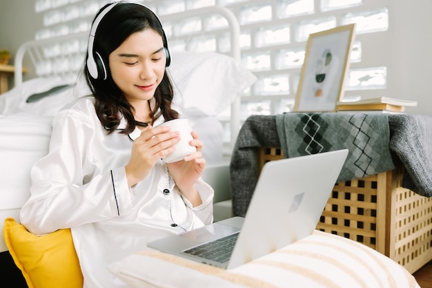 Beautiful young Asian woman sitting next to bed using laptop and breakfast in the morning at home