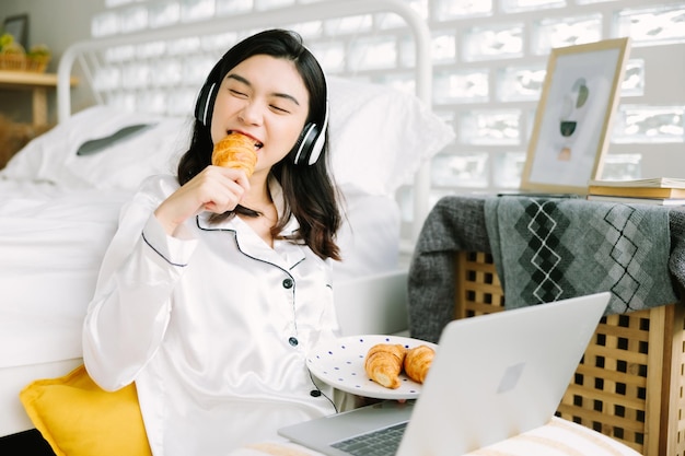 Beautiful young Asian woman sitting next to bed using laptop and breakfast in the morning at home
