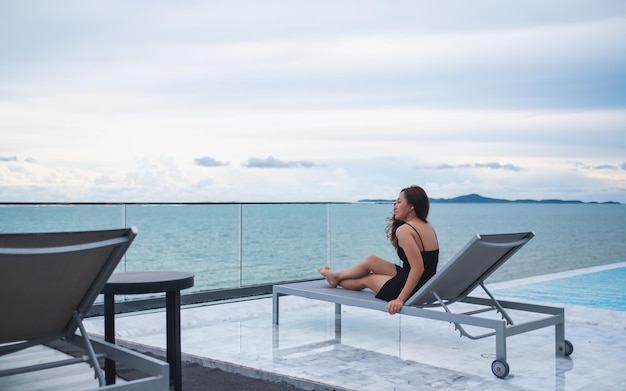 屋上プールの隣に座って、海と青空の景色を見ている美しい若いアジアの女性