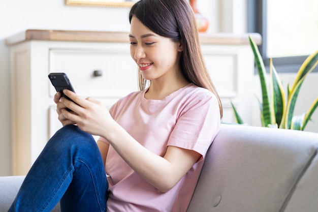 Beautiful young Asian woman sitting on sofa and using phone to text