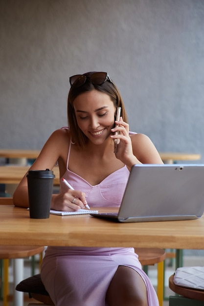 Beautiful young asian woman sitting with laptop at cafe calling by smartphone