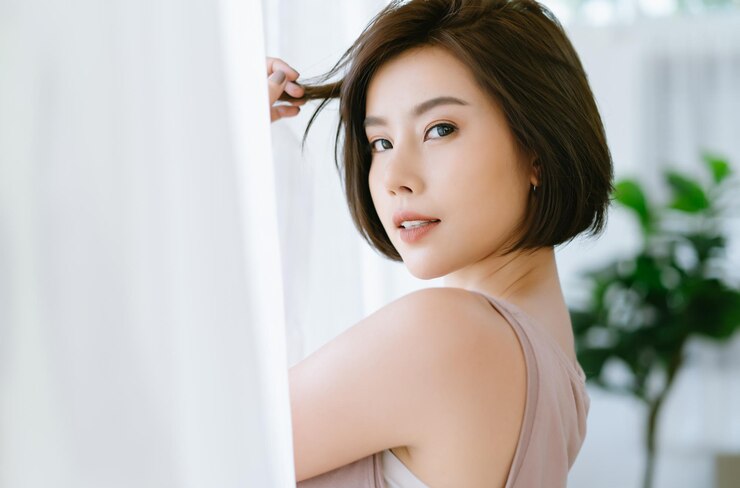 Photo 창 흰색 커튼 근처에 서서 카메라를 보고 미소를 짓는 아름다운 젊은 아시아 여성