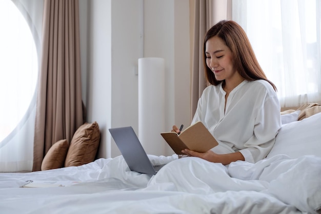Красивая молодая азиатка, использующая портативный компьютер и работающая над документами, сидя дома на кровати