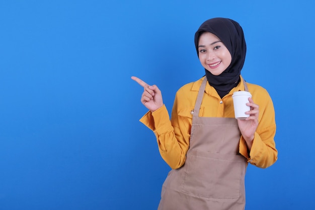 Beautiful young asian woman wear apron holding a glass of coffee and pointing left side