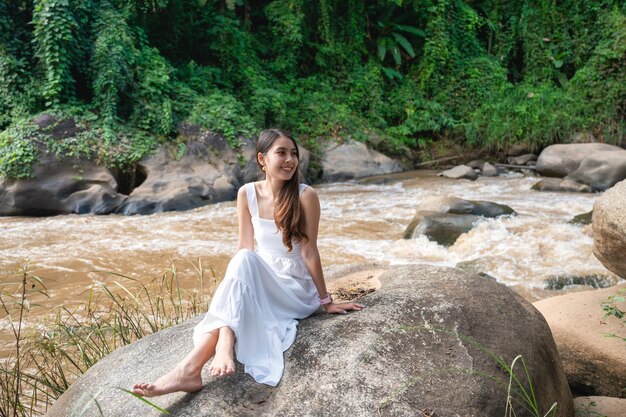 휴가철 계곡의 강변에서 휴식을 취하는 하얀 드레스를 입은 아름다운 젊은 아시아 여성. 여가 활동, 레크리에이션 추구