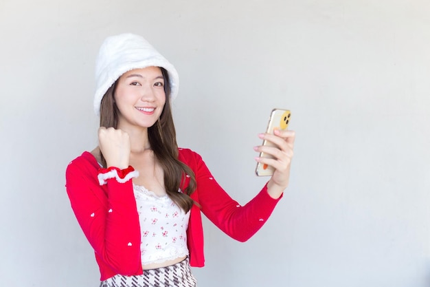 Beautiful young Asian woman who wears a red coat and white hat as a Santy girl holds a smartphone